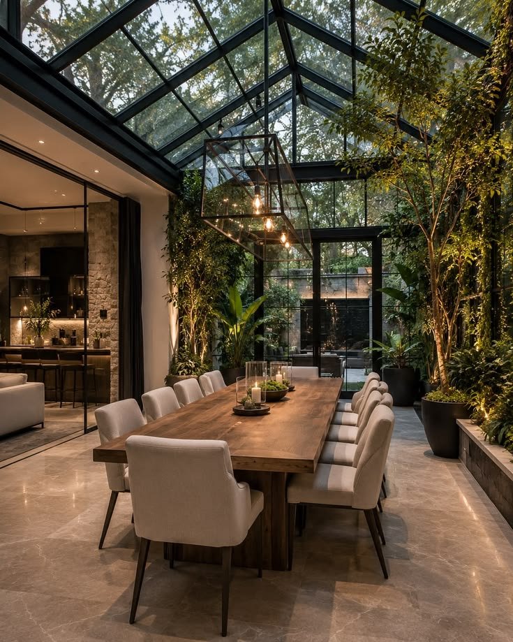 Glass-roofed dining room with a long timber table, cream chairs and trees growing beside it
