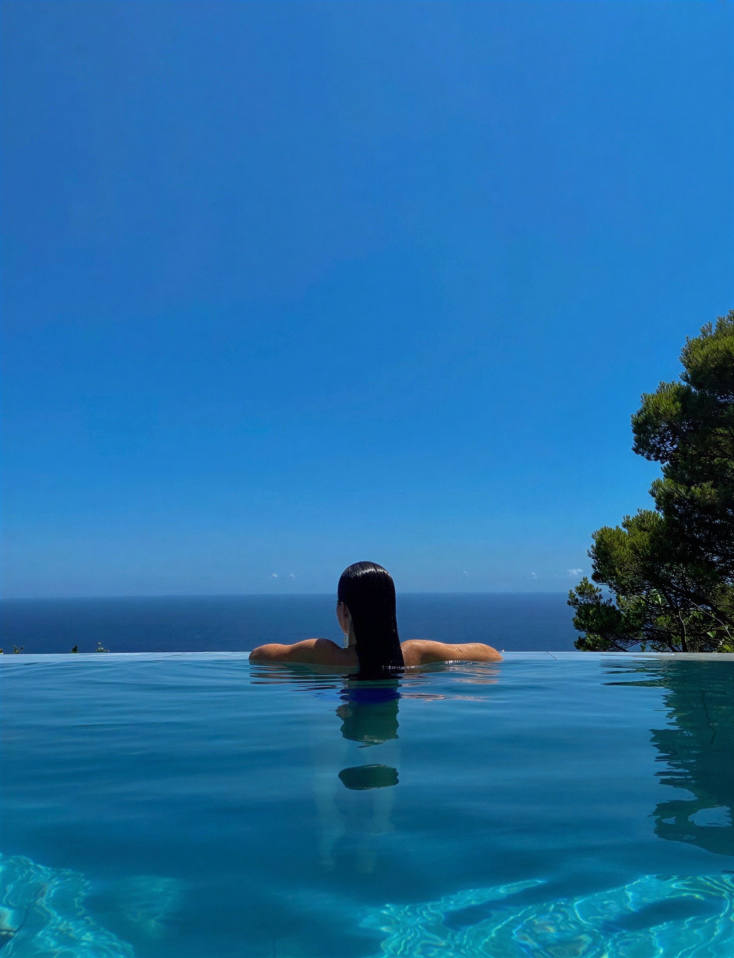 A woman seen from behind resting at the edge of an infinity pool, looking out over the sea